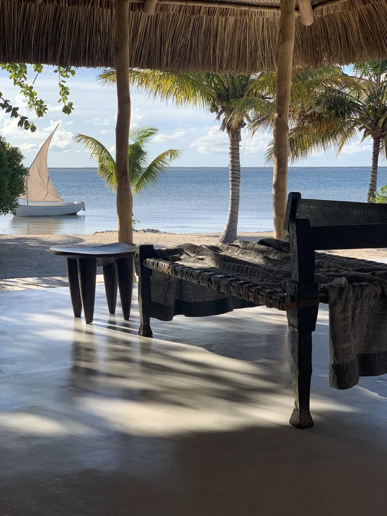 Wooden day bed with view of beach and dhow on the ocean.